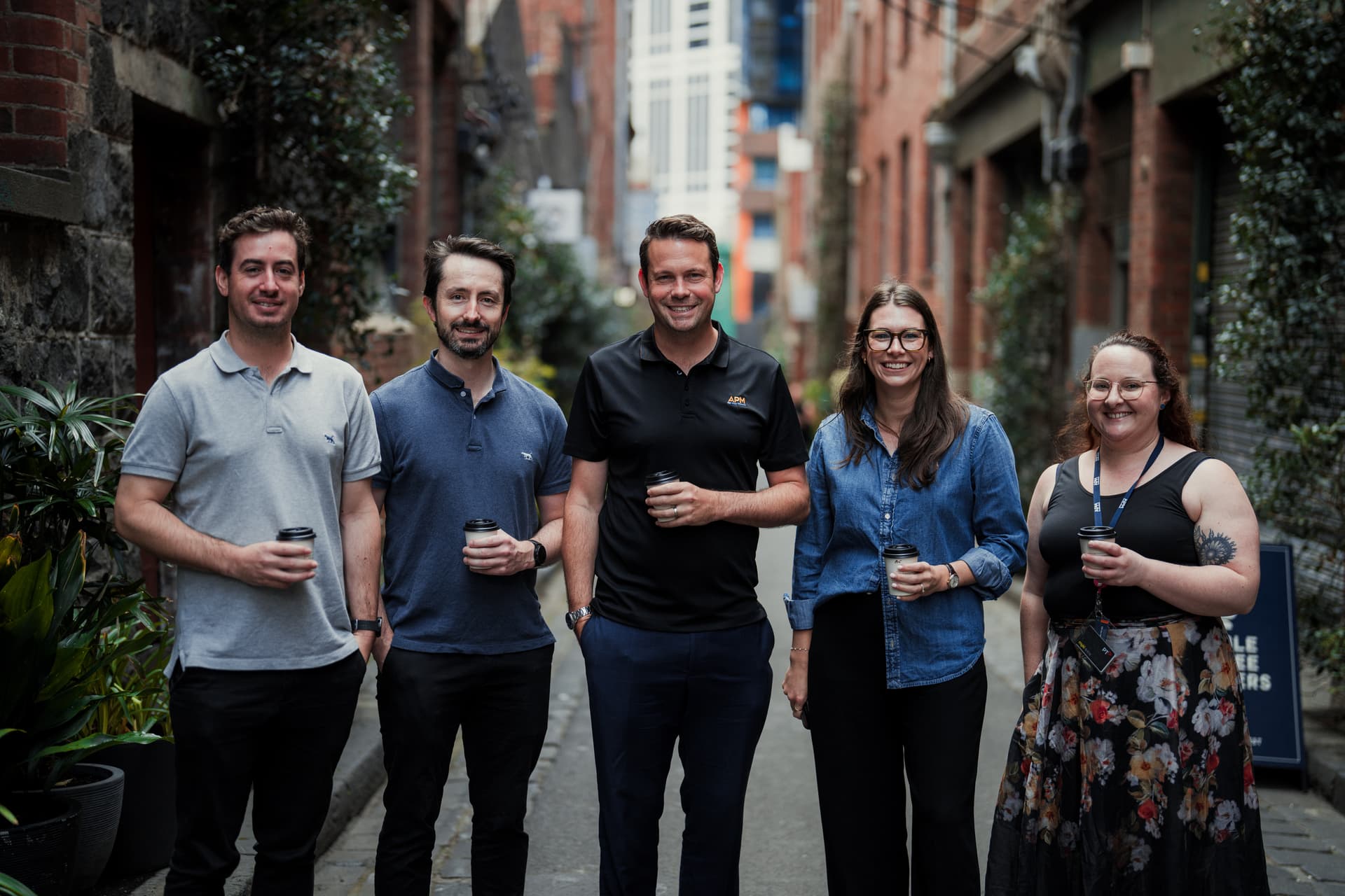 Five APM WorkCare team members standing casually in a line holding coffees