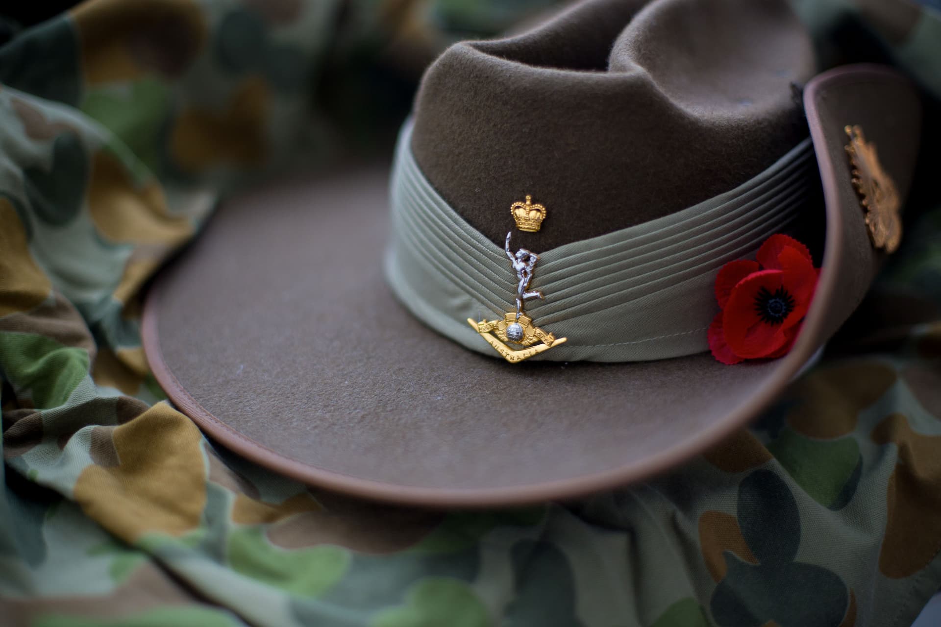 Australian Army slouch hat resting on camouflage fabric, featuring military insignia and a red remembrance poppy.