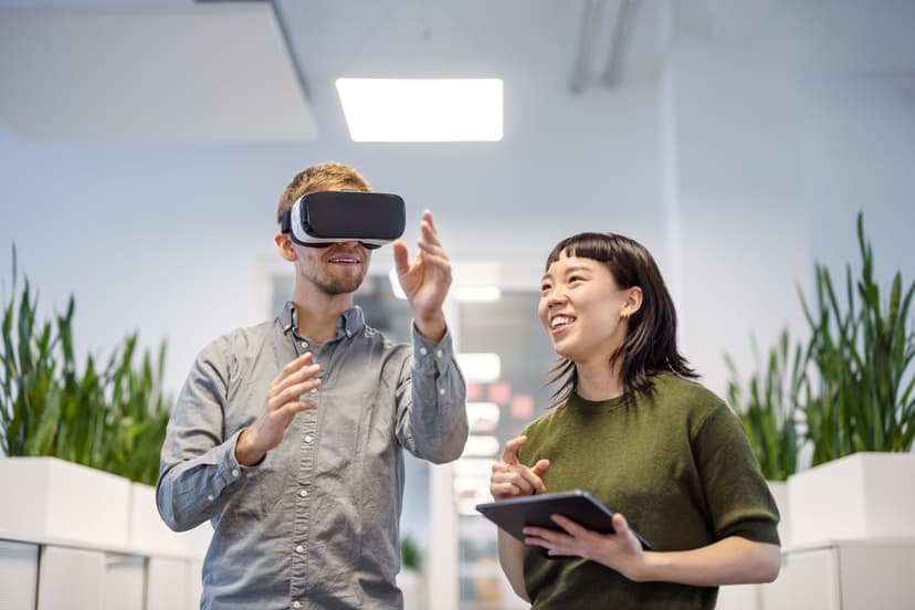 Two individuals in a work a setting. One has a VR headset, the other holds a tablet while instructing the VR headset individual
