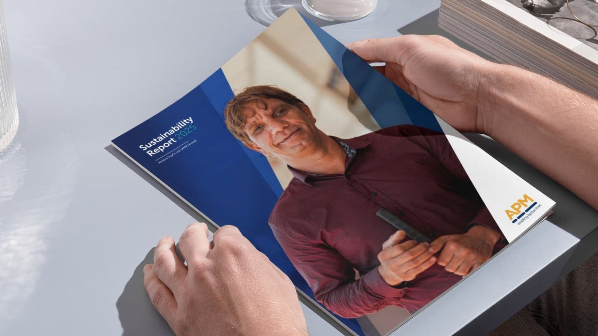 A person holding a printed copy of the APM Sustainability Report 2025, showing the cover design and branding.