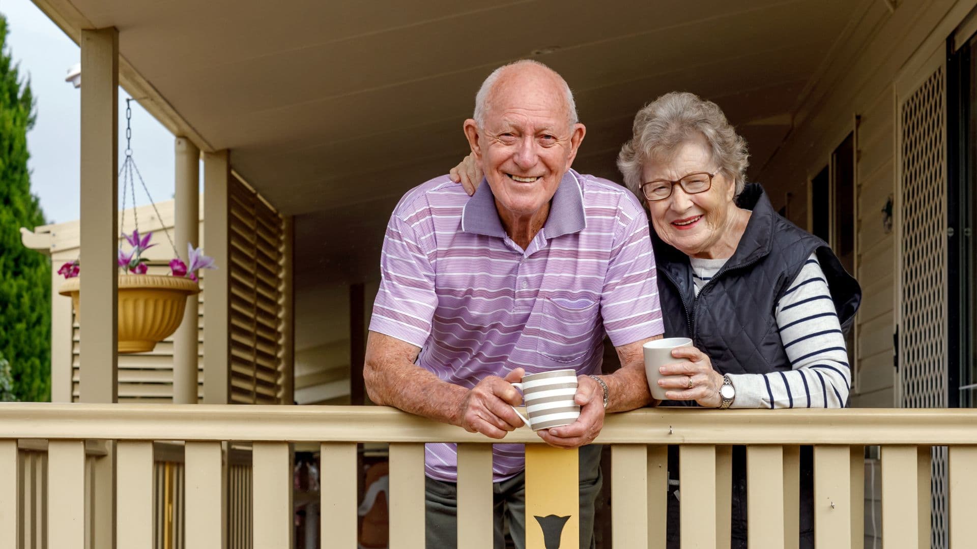 Two older adults leaning on a porch railing with drinks, illustrating ageing at home and lifestyle factors considered during aged care assessments.