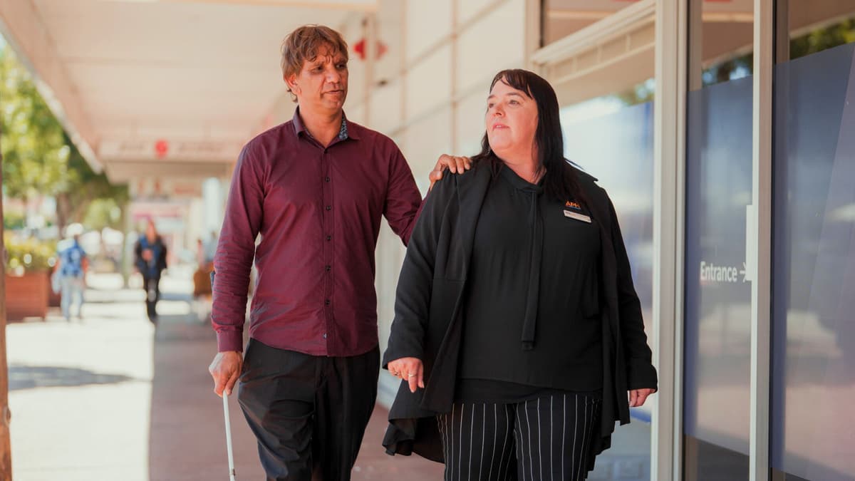 Jamie, a First Nations NDIS participant, walks with a cane and a hand resting on the shoulder of Mykala from APM Communities.