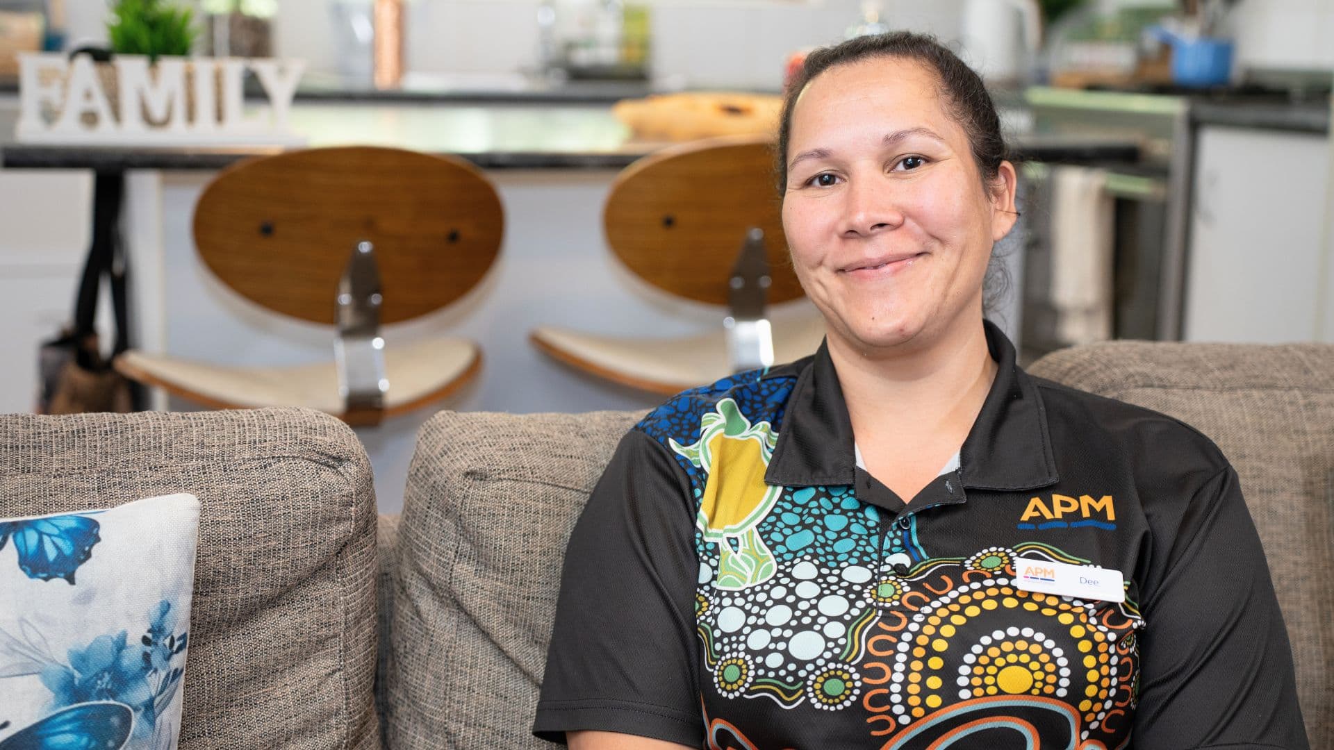 APM team member Dee in a First Nations top on a sofa at home