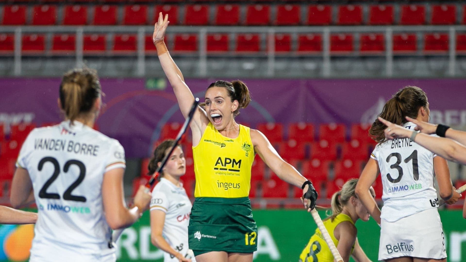Australian women’s hockey team player in yellow and green uniform raising an arm in celebration during a match against opponents in white uniforms. APM and Ingeus logos are visible on the uniform. Red stadium seats and purple banners in the background.