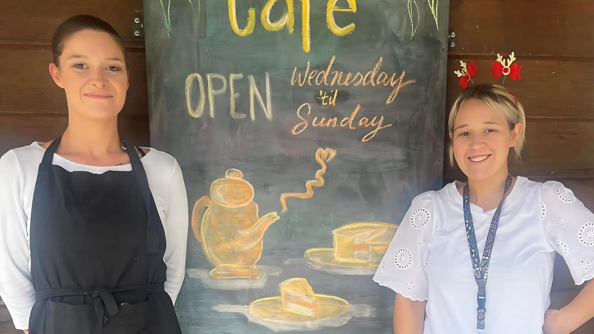 Briannon in a black apron next to a cafe chalk board and her APM planner