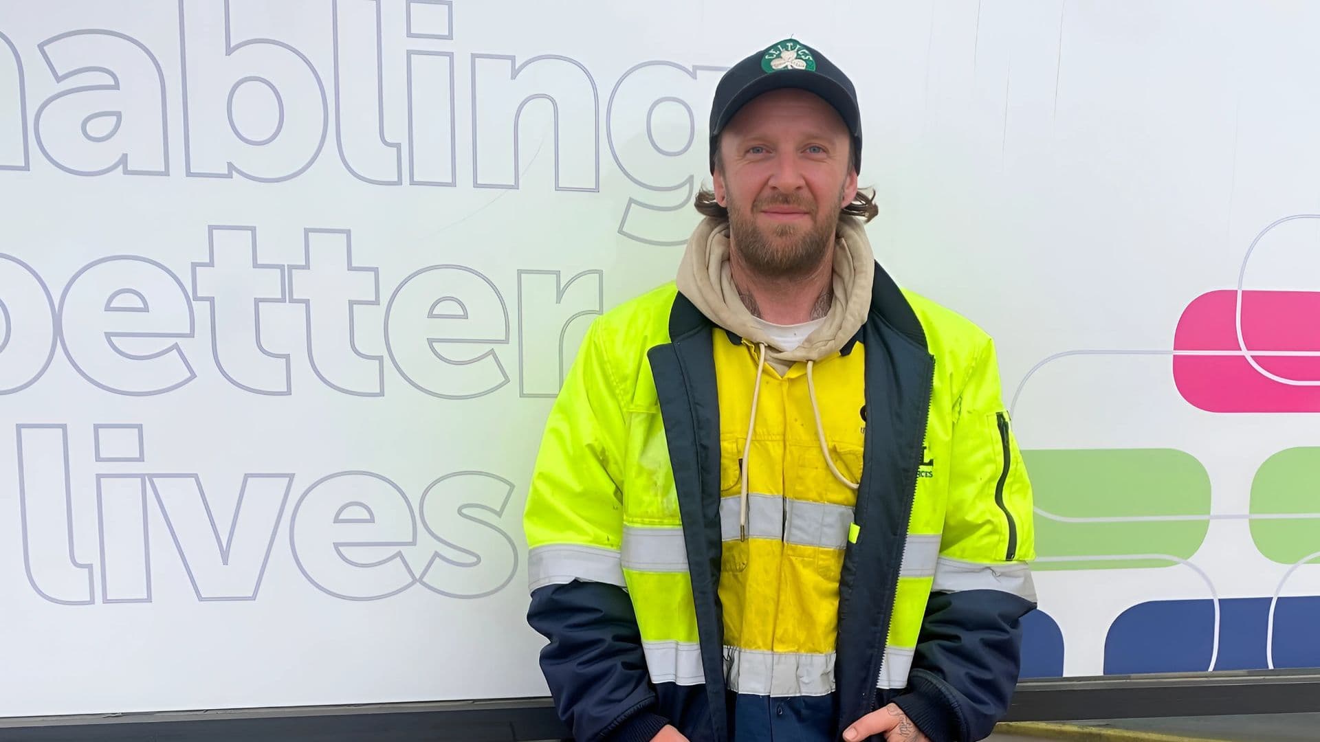 Myran a DES participant, stands outside an APM branch in a high-vis jacket and a green Celtics cap.