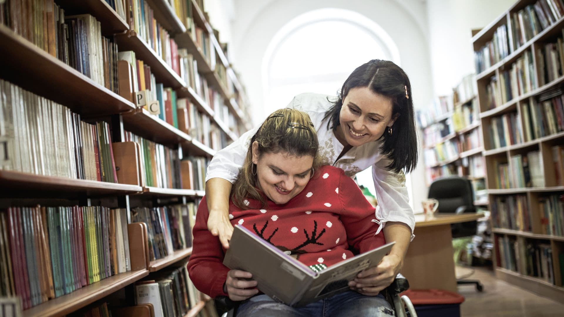 A woman smiles reading a book with a female carer in a library