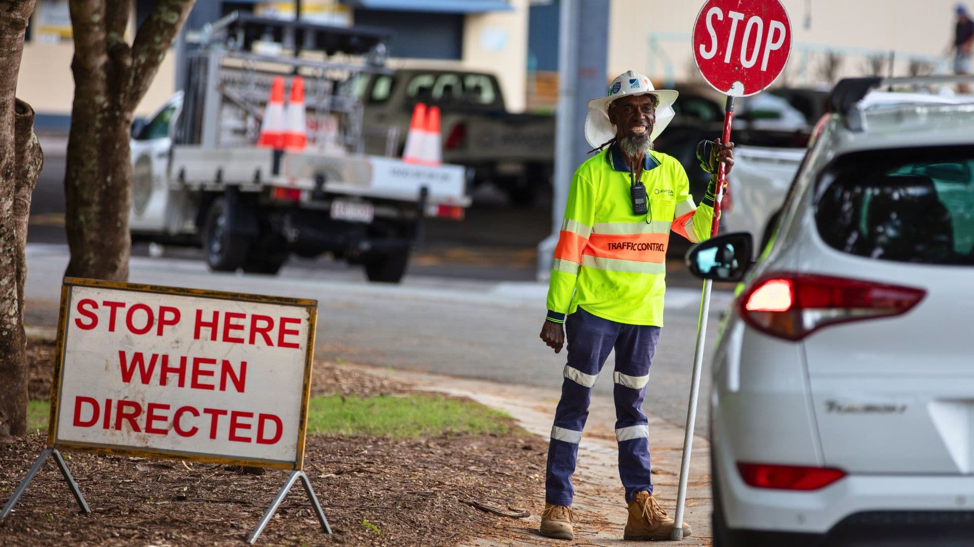 Workforce Australia participant Adam directs traffic holding a stop sign, standing next to a "Stop Here When Directed" sign, with vehicles and traffic control equipment visible in the background.