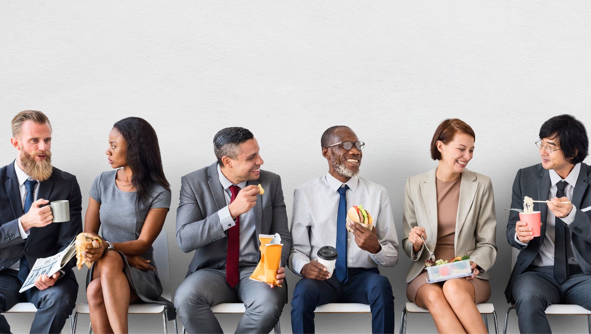 A row of professional office workers sitting against a wall eating various lunches