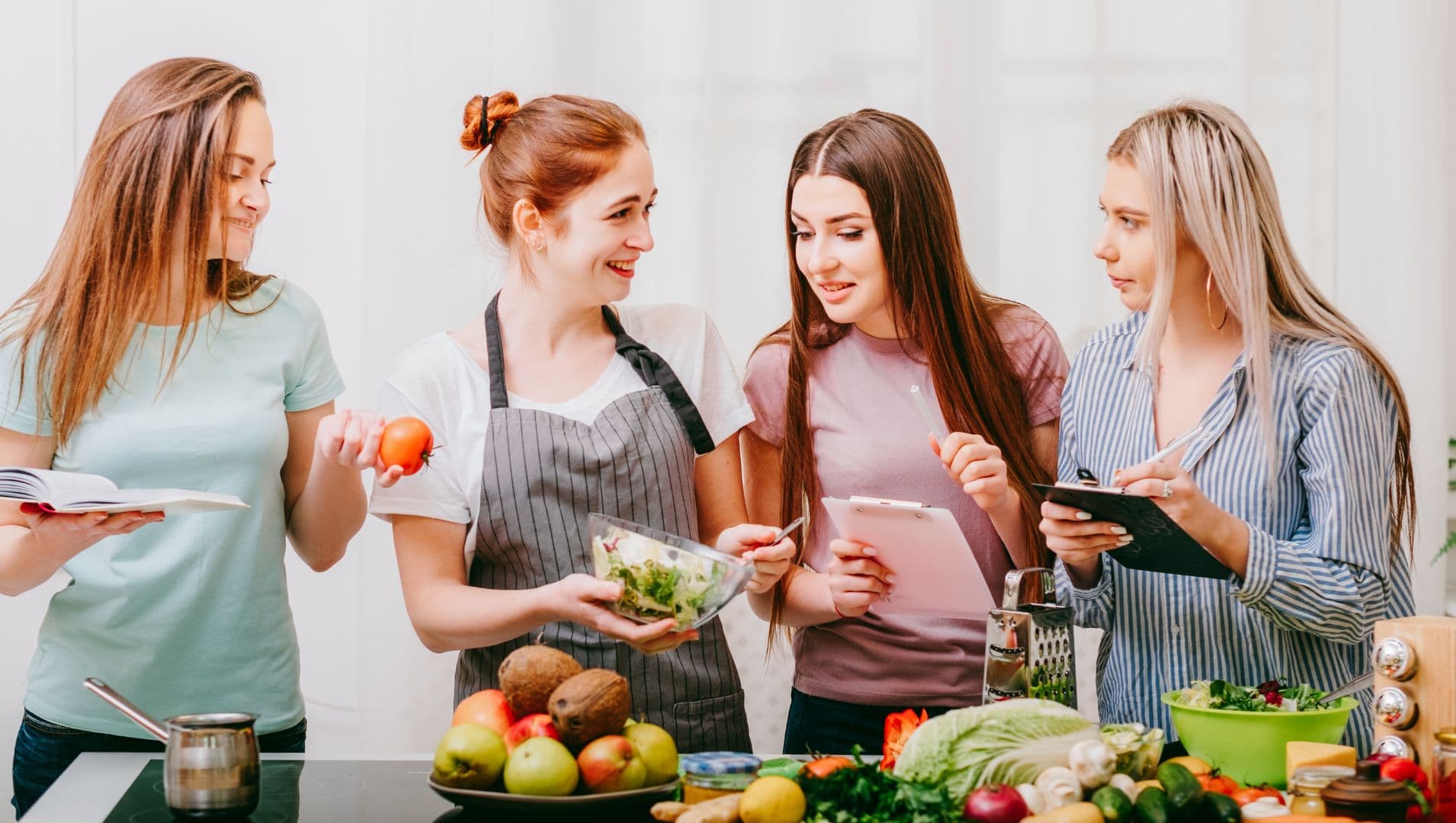 A woman in an apron discusses healthy food with three other women next to a counter of fresh healthy food.