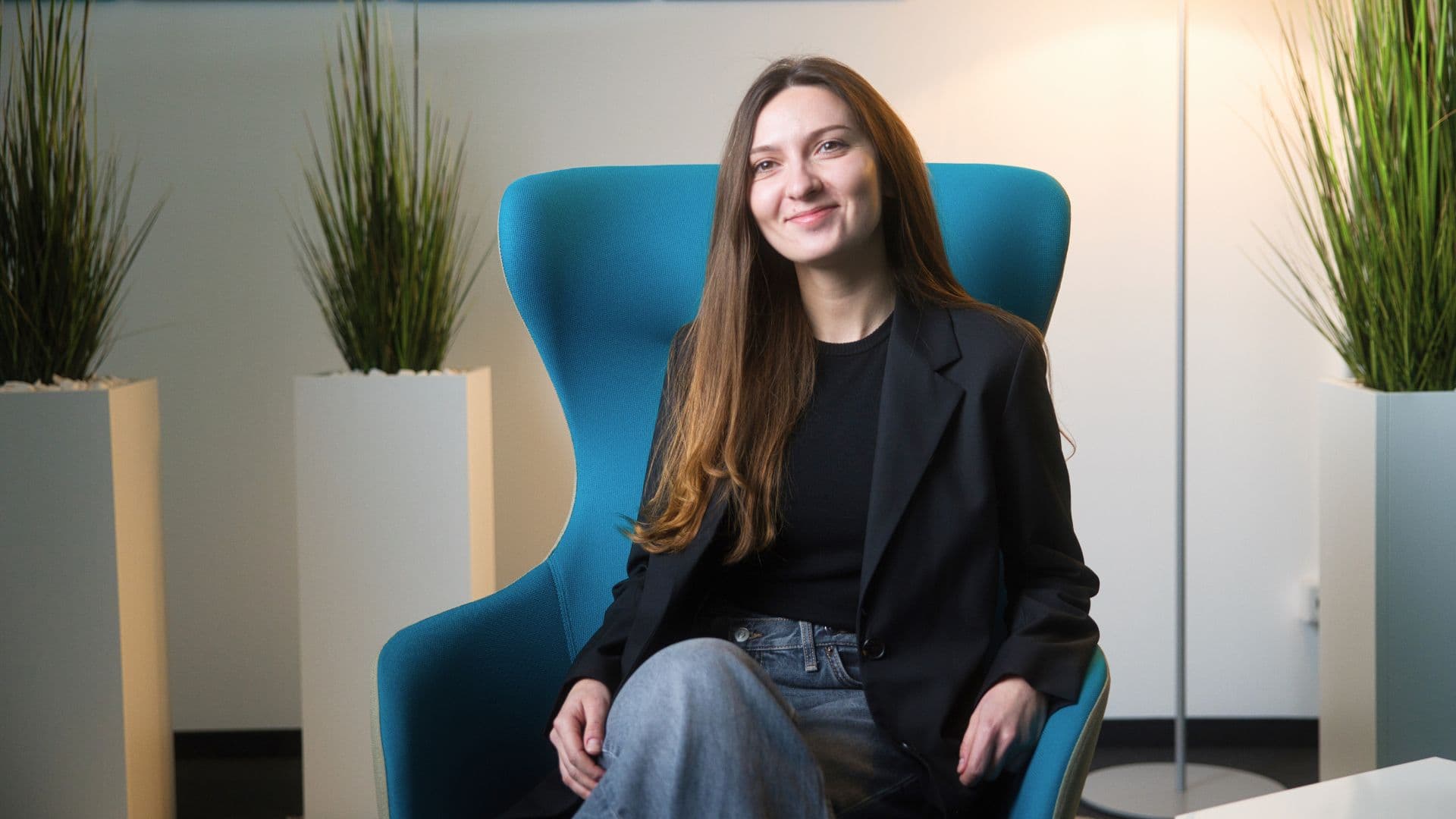 Person sitting in a blue chair with a smile, wearing a black blazer and jeans, flanked by potted plants in a modern office setting.