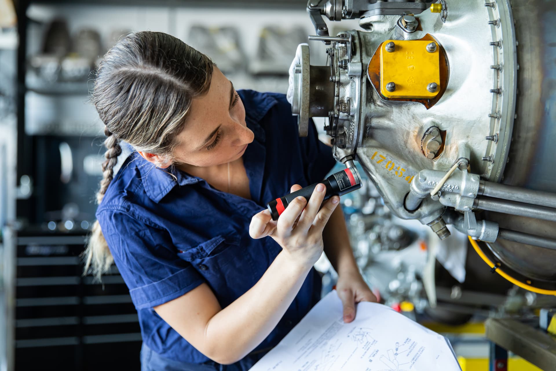 Apprentice inspecting an aircraft engine component with a torch while reviewing technical drawings in a maintenance workshop.