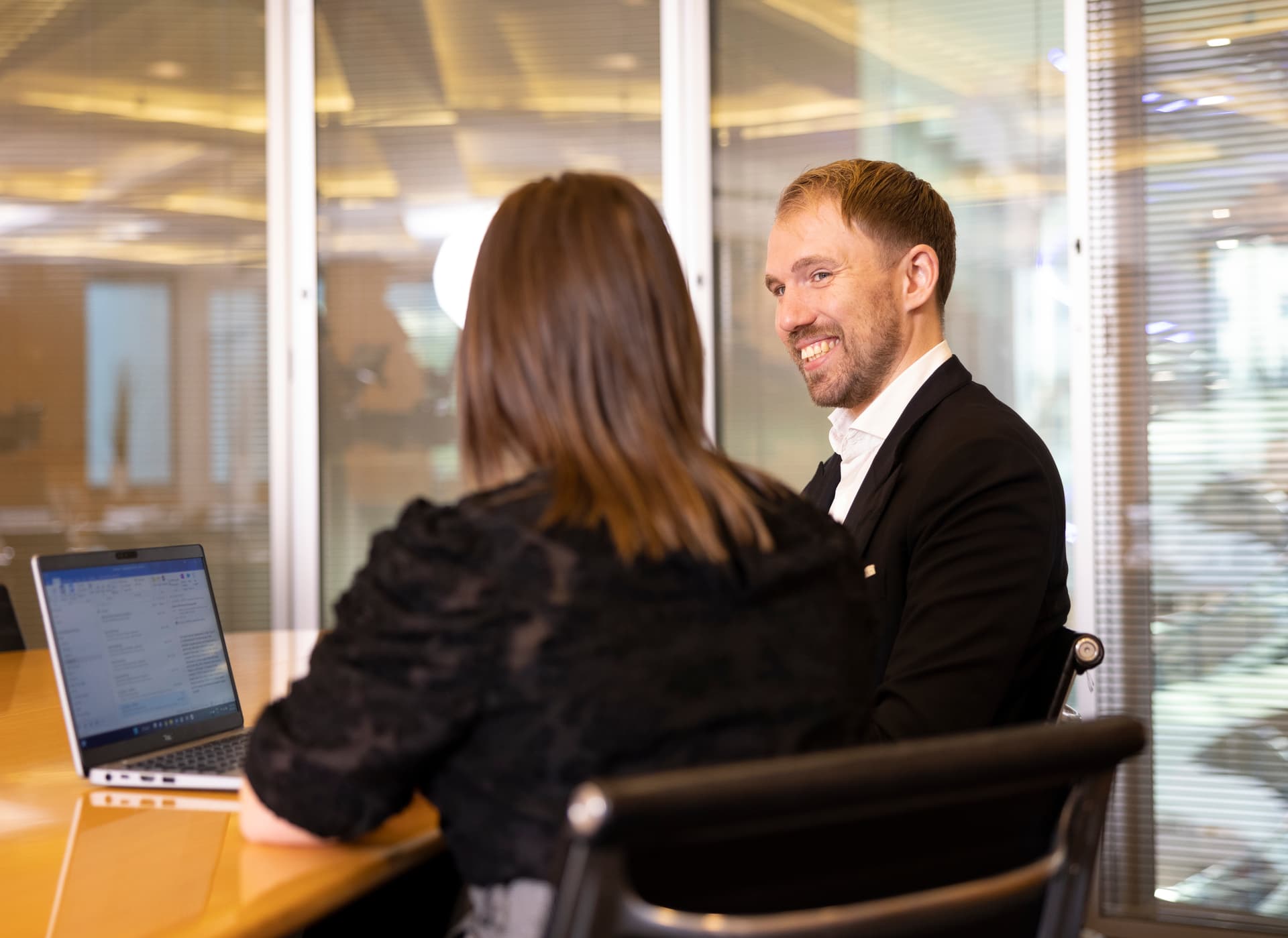 Two professionals seated across a meeting table in a glass-walled office meeting room. An open laptop is visible on the table as they engage in a workplace discussion. The space features modern furnishings, large windows, and bright natural lighting.