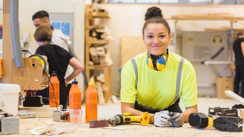 Apprentice working at a woodworking bench in a trade workshop, wearing safety gear and surrounded by tools and equipment