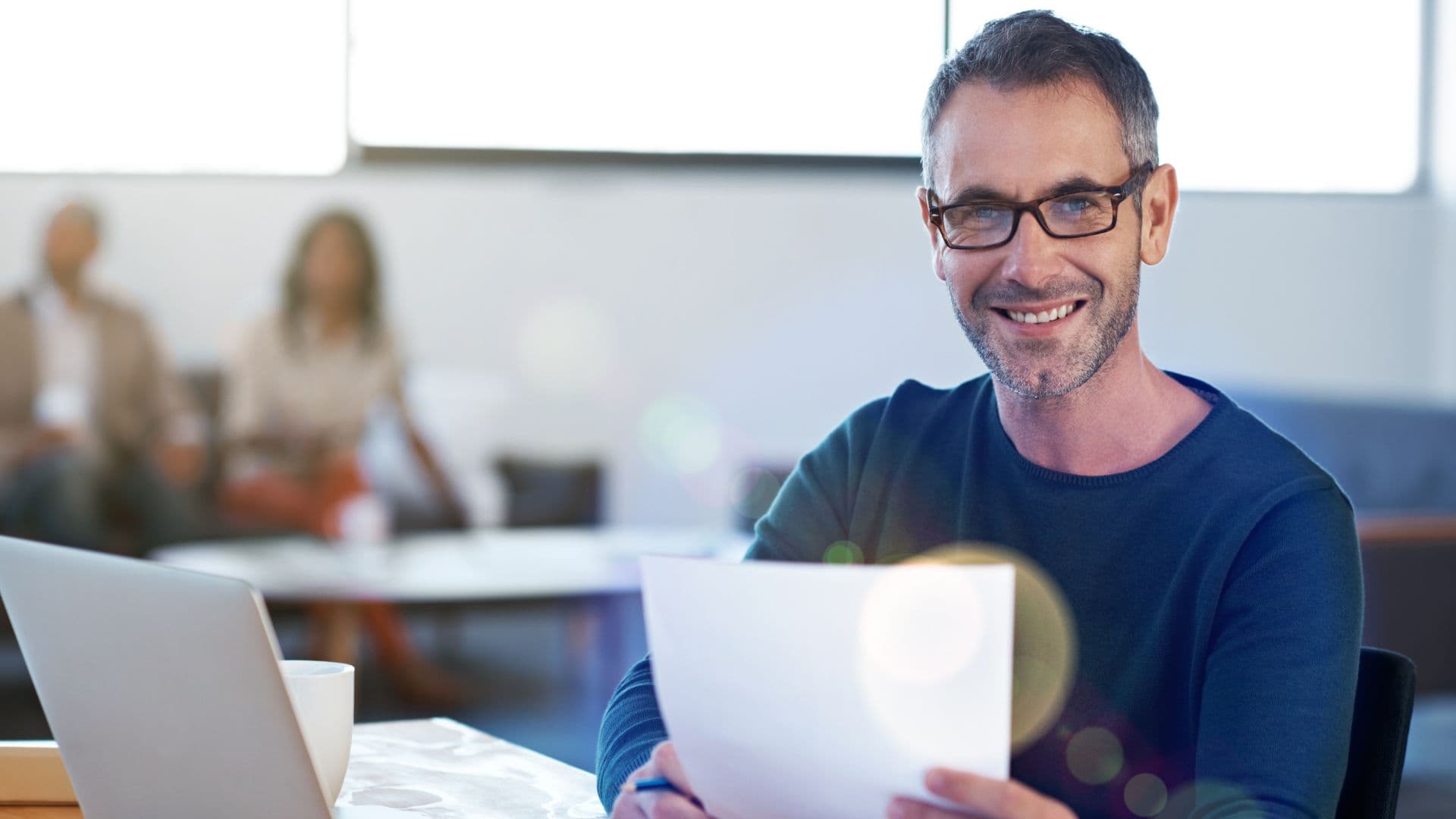 A man sitting at a desk smiling while holding a sheet of paper