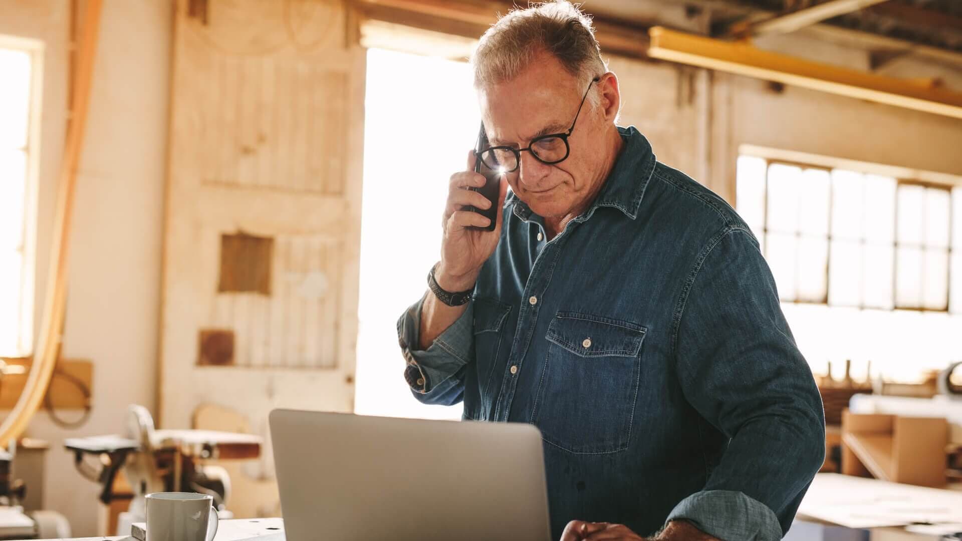 A man standing in front of a laptop and talking on the phone in his workshop