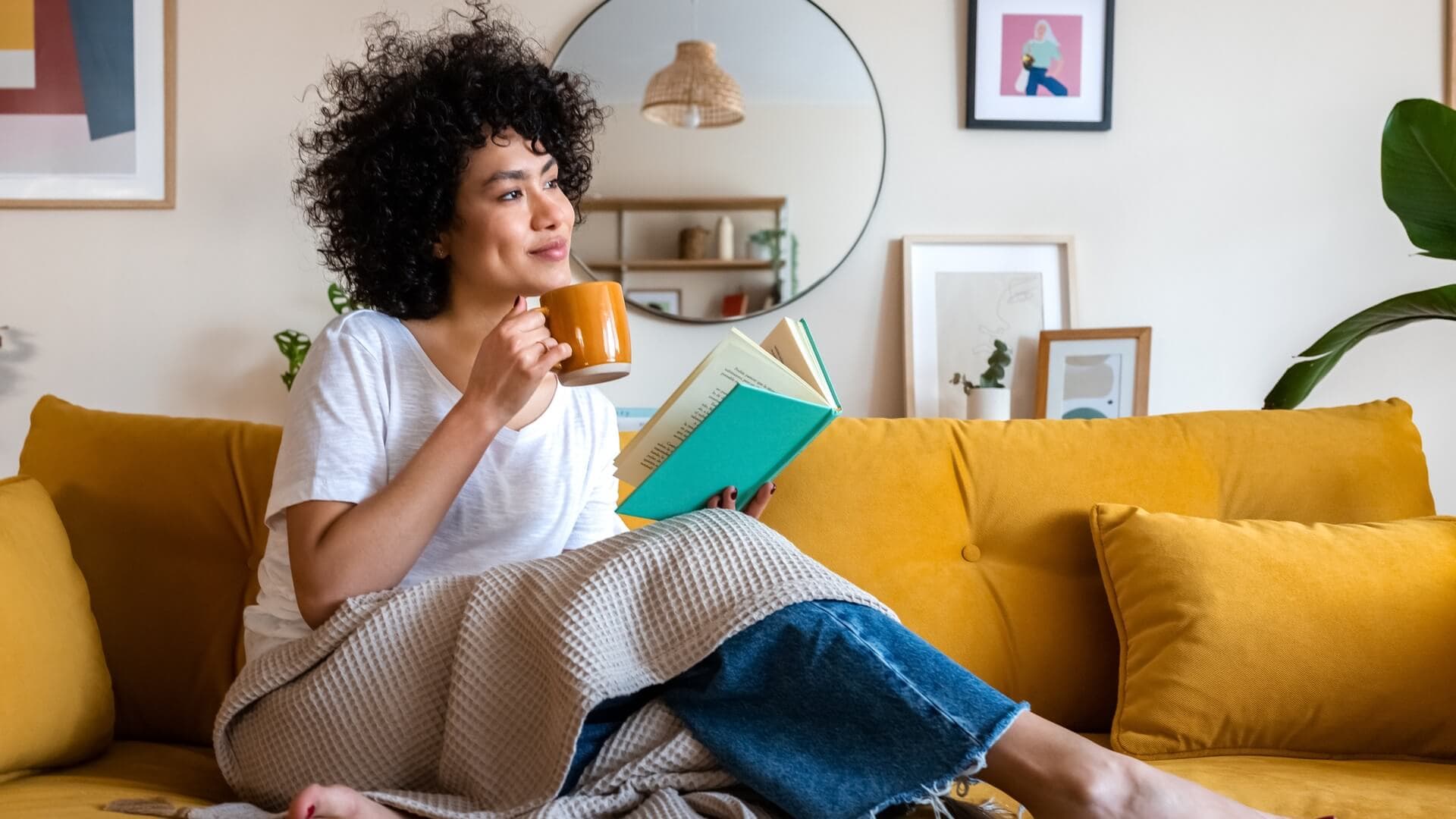 A woman reading a book and drinking a cup of coffee on a couch
