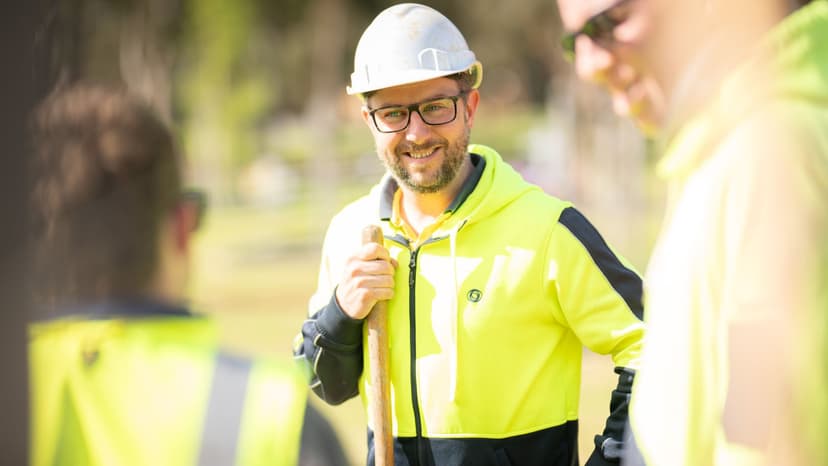 A construction worker wearing a hard hat and high-visibility jacket smiles while holding a measuring tape, with two colleagues partially visible in the foreground.